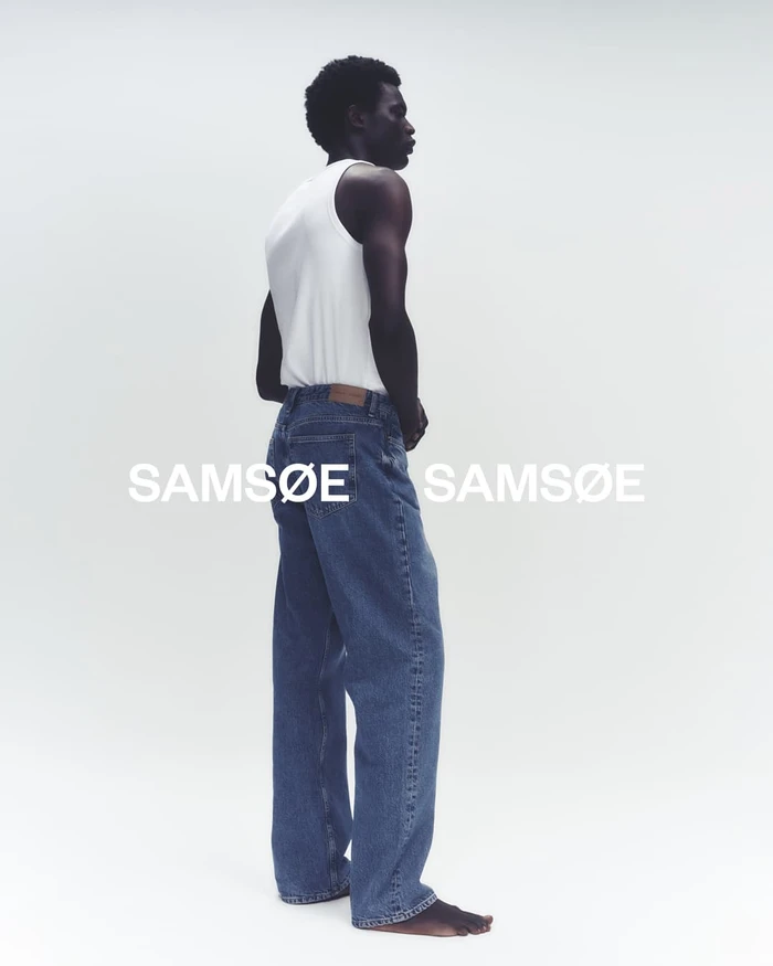 Black man wearing a white tank top and blue denim jeans posing against a white background for Samsøe Samsøe
