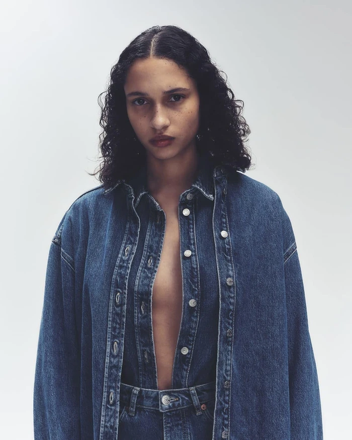 Model with dark curly hair wearing an unbuttoned denim shirt and matching denim jeans against a plain light grey background.