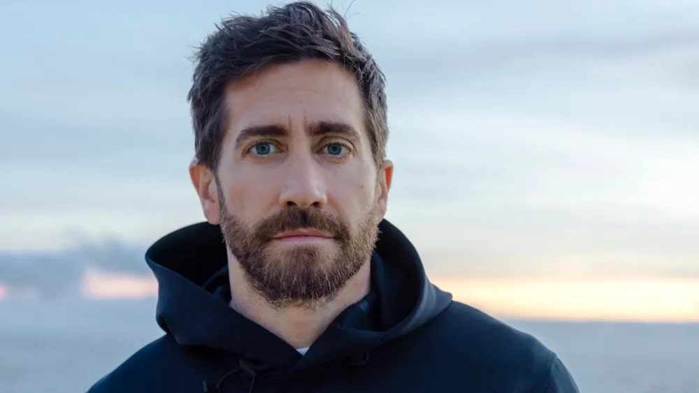 Headshot of an adult male with a beard, wearing a hooded sweatshirt, against a blurred background of the sky and possibly a body of water.