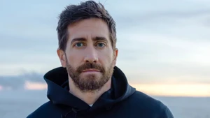 Close-up portrait of actor Jake Gyllenhaal wearing a black hooded sweatshirt against a soft sunset sky backdrop.