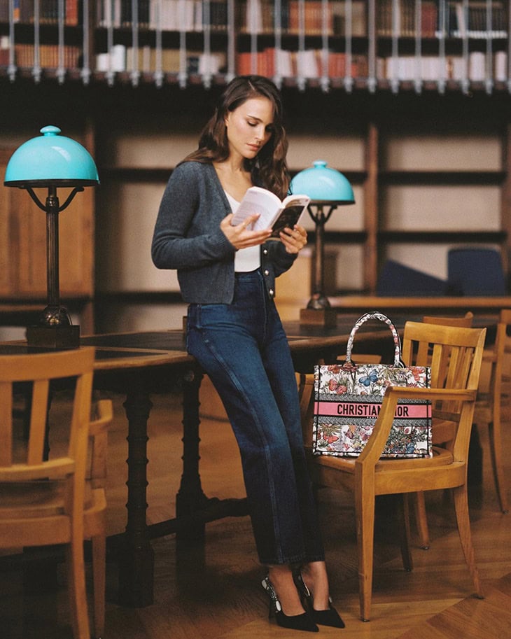 A woman in jeans and a cardigan is reading in a library, with a branded bag nearby, under warm light, with bookshelves in the background.