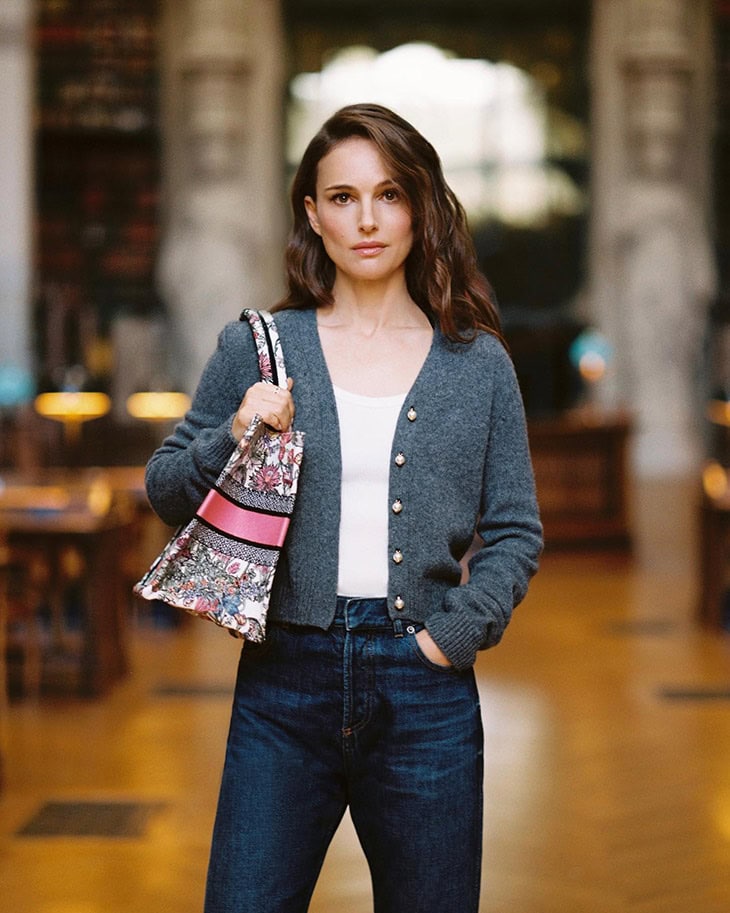 Portrait of Natalie Portman in a library, holding a patterned tote bag, wearing a gray cardigan and jeans, posing indoors.