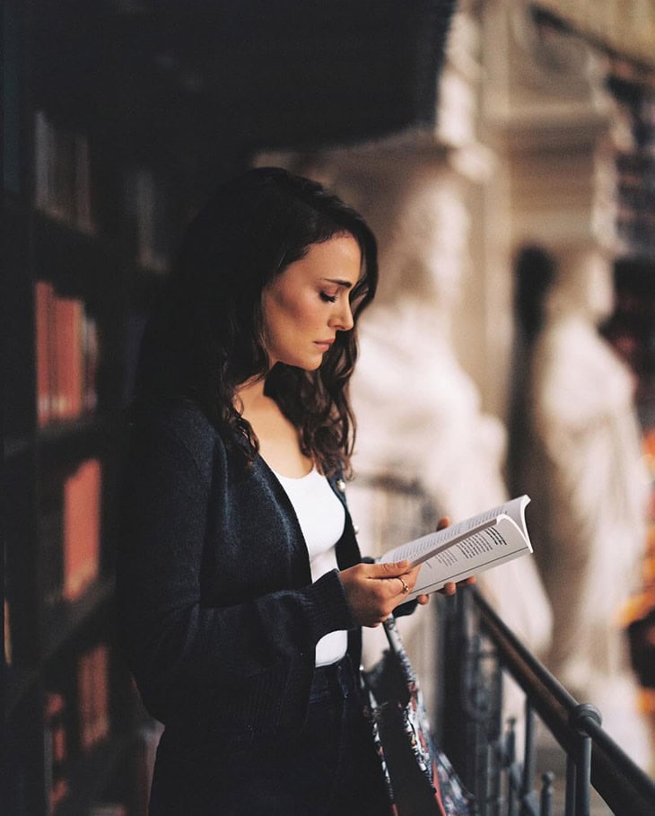 Woman reading a book in a library, wearing a dark cardigan and a white top, standing by a railing, statues in the background.