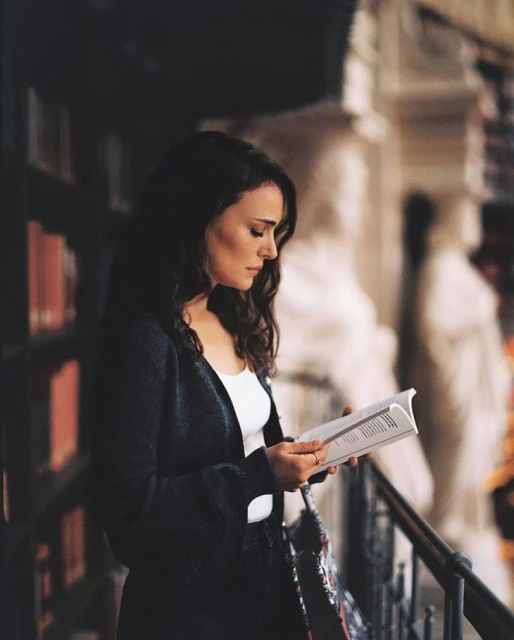 Woman reading a book in a library, wearing a dark cardigan and a white top, standing by a railing, statues in the background.