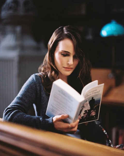 Young woman with long brown hair sits indoors reading a book with a figure on the cover, near a wood banister and muted blue lamp.