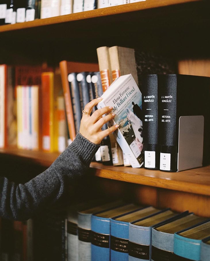 Woman's hand pulling Elena Ferrante book from a wooden library bookshelf stocked with an assortment of books in a warm indoor setting.