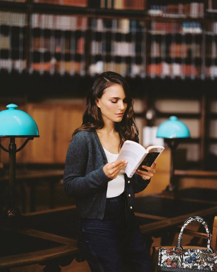 Portrait of a woman reading in a library. She is wearing a cardigan. Bookshelves and a bag are in the background.
