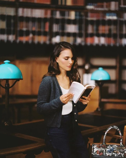 Portrait of a woman reading in a library. She is wearing a cardigan. Bookshelves and a bag are in the background.