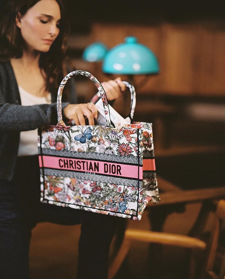 Adult woman holding a Christian Dior floral tote bag in an indoor setting with warm lighting, showcasing fashion and luxury.