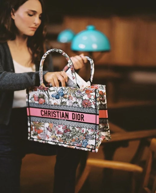 Adult woman holding a Christian Dior floral tote bag in an indoor setting with warm lighting, showcasing fashion and luxury.