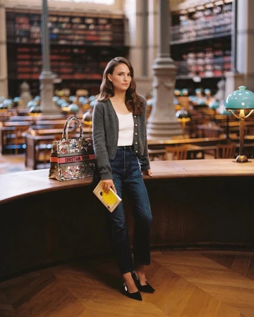 Adult woman poses in a library, wearing jeans, a cardigan, and holding a book with a Christian Dior bag on a desk.