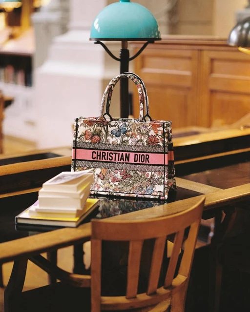 Christian Dior tote bag with floral pattern on a wood desk in a library-like space with a turquoise lamp and stacked books.