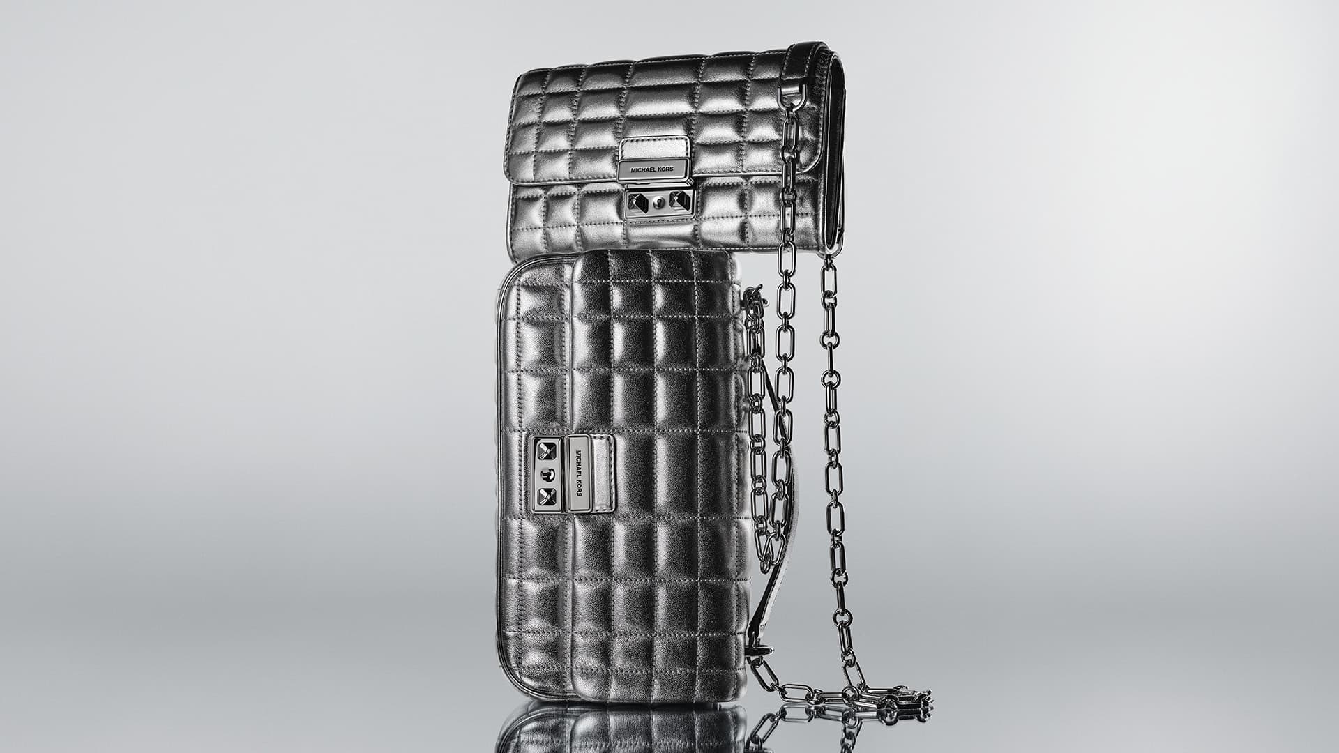 Two metallic silver quilted Michael Kors handbags, one stacked on top of the other, with a silver chain against a gradient grey background.