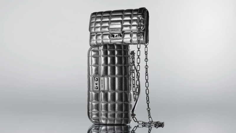 Two metallic silver quilted Michael Kors handbags, one stacked on top of the other, with a silver chain against a gradient grey background.