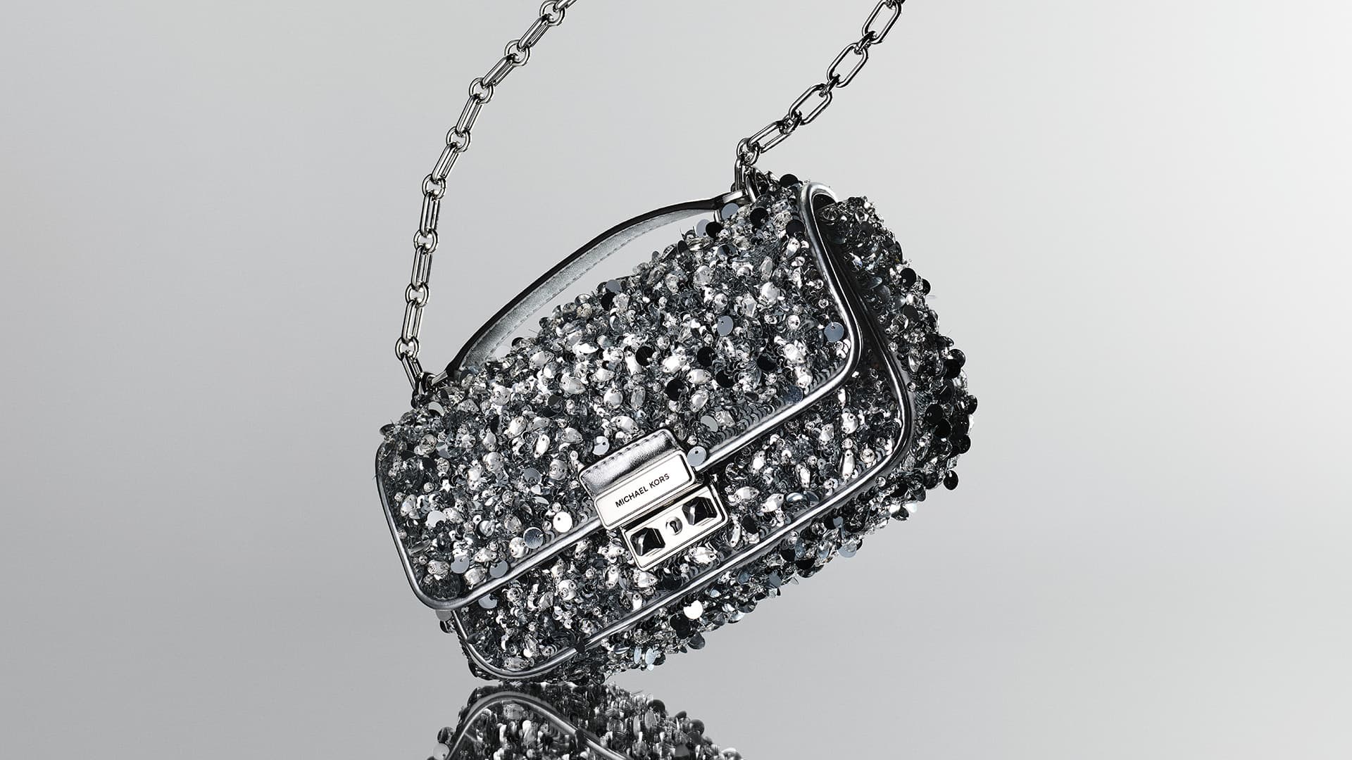 Silver Michael Kors purse with a metal chain strap and encrusted with small silver embellishments, studio shot on a grey background.