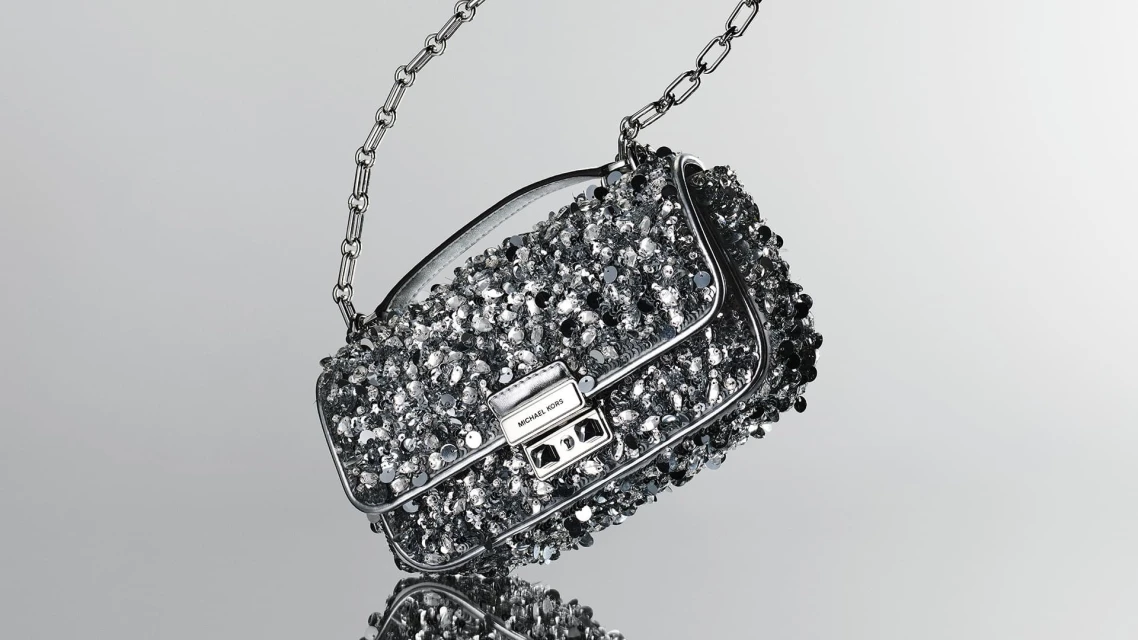 Silver Michael Kors purse with a metal chain strap and encrusted with small silver embellishments, studio shot on a grey background.