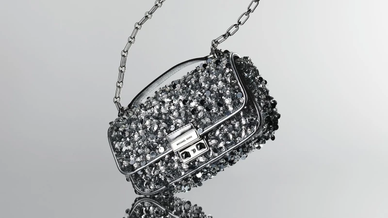 Silver Michael Kors purse with a metal chain strap and encrusted with small silver embellishments, studio shot on a grey background.
