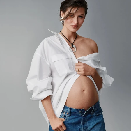 Pregnant woman in unbuttoned white shirt and blue jeans poses in a studio with a gray backdrop, showcasing maternity fashion.