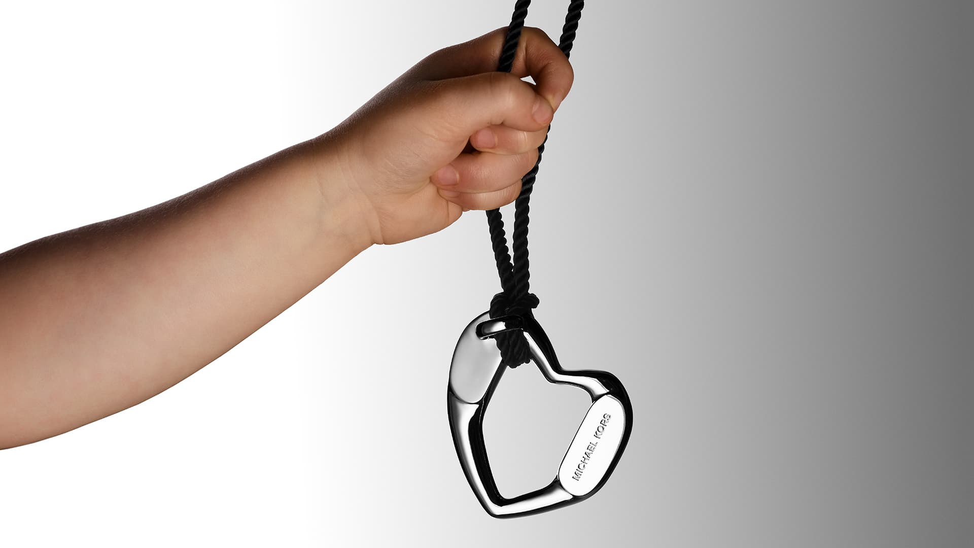 Hand holds a shiny heart-shaped Michael Kors pendant necklace against a gray-white gradient background, showcasing jewelry fashion accessory.