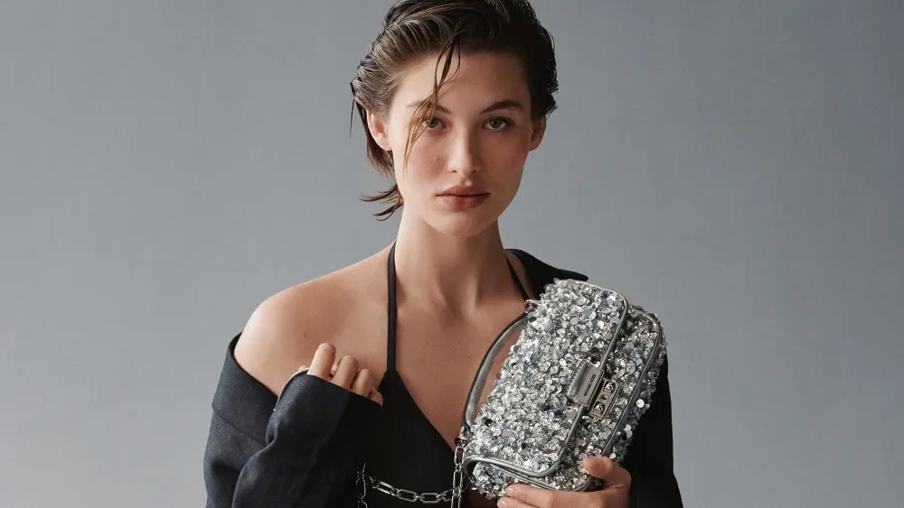 Close-up of a model holding a silver sequin purse, wearing a black halter top against a grey backdrop in a studio lighting setup.