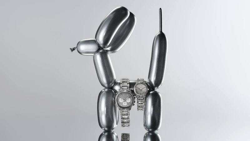 Two silver wristwatches positioned on a reflective surface in front of a balloon dog sculpture on a gray gradient background.