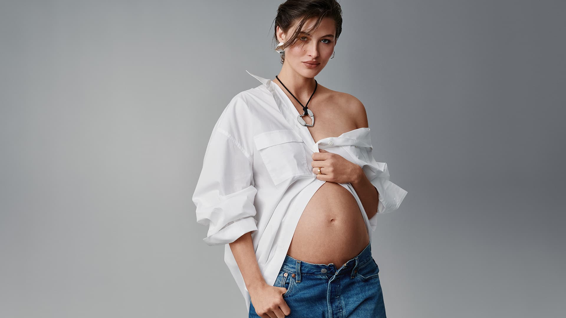 A pregnant woman poses in a studio wearing a white shirt and blue jeans, looking directly at the camera, with a grey backdrop.