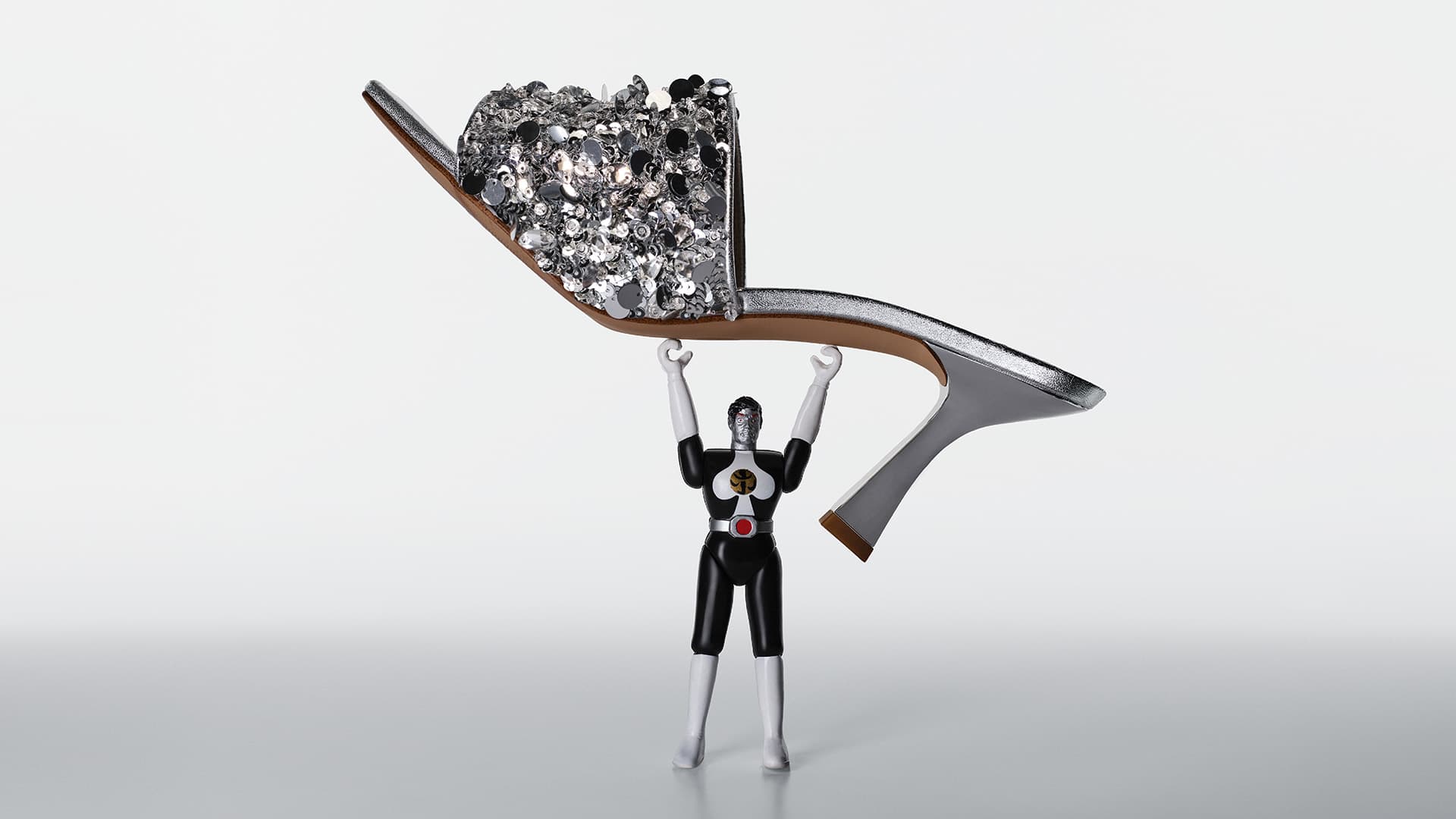 Toy figure holds up a silver high-heeled shoe decorated with silver sequins against a bright white background in a studio shot.