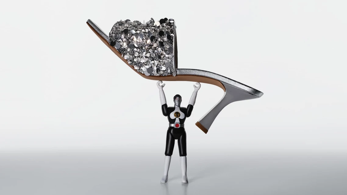 Toy figure holds up a silver high-heeled shoe decorated with silver sequins against a bright white background in a studio shot.
