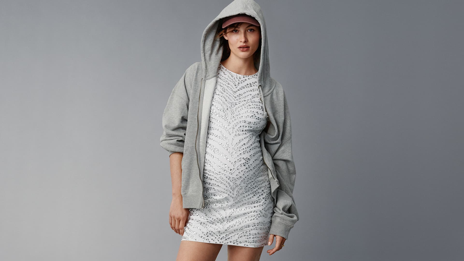 A young woman poses in a studio wearing a printed mini dress, grey hoodie, and pink baseball cap, gazing directly at the camera.