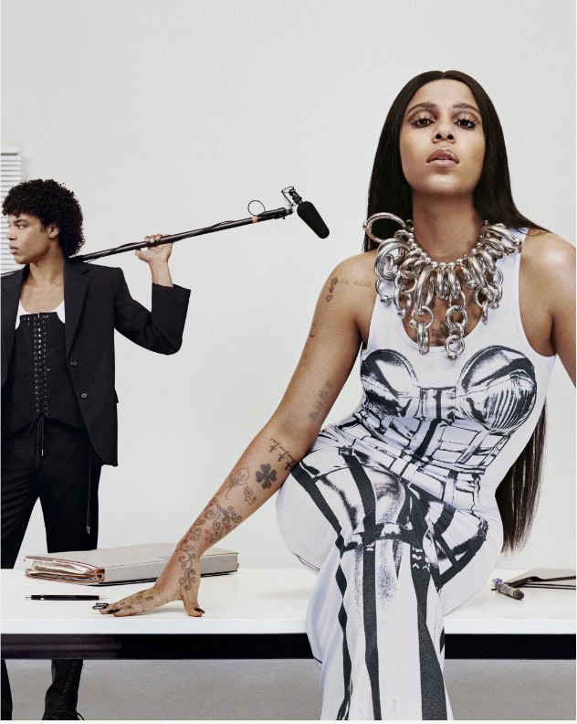 Stylish women and man in fashion shoot. Woman in patterned top sits on desk. Man in suit holds microphone. Minimal background.