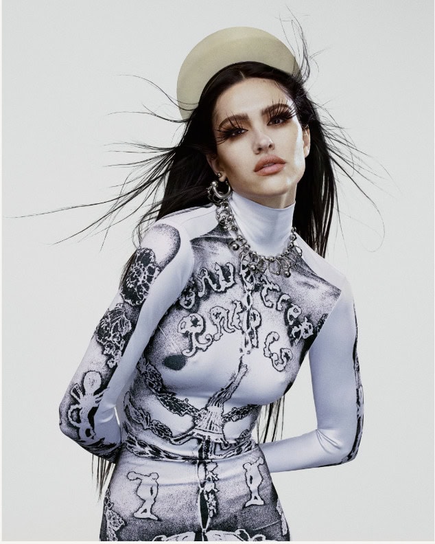 Portrait of woman with long dark hair, halo, heavy eyelashes, statement jewelry wearing a printed white bodysuit in a photo studio.