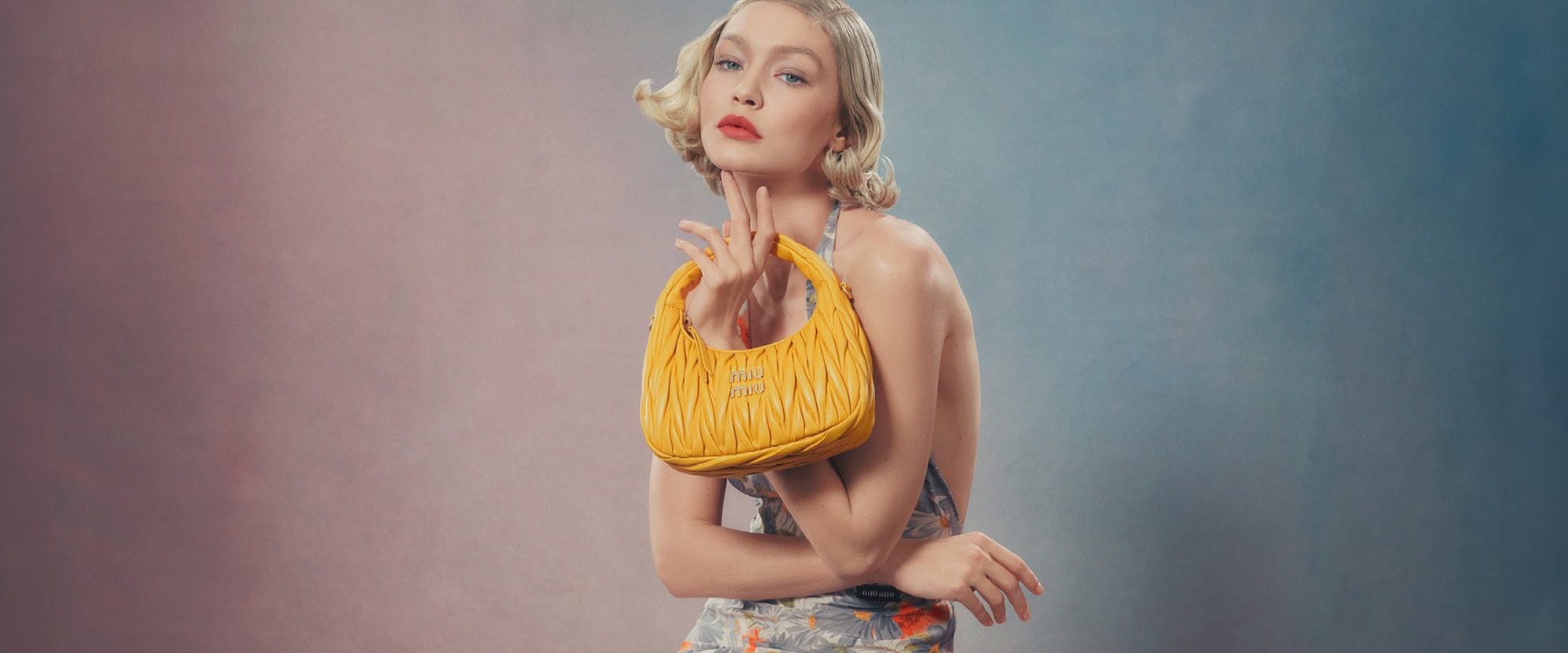 Model Gigi Hadid holding a yellow quilted handbag against a gradient studio background for Miu Miu spring 2024 campaign