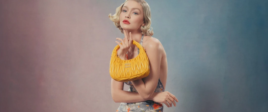 Model Gigi Hadid holding a yellow quilted handbag against a gradient studio background for Miu Miu spring 2024 campaign
