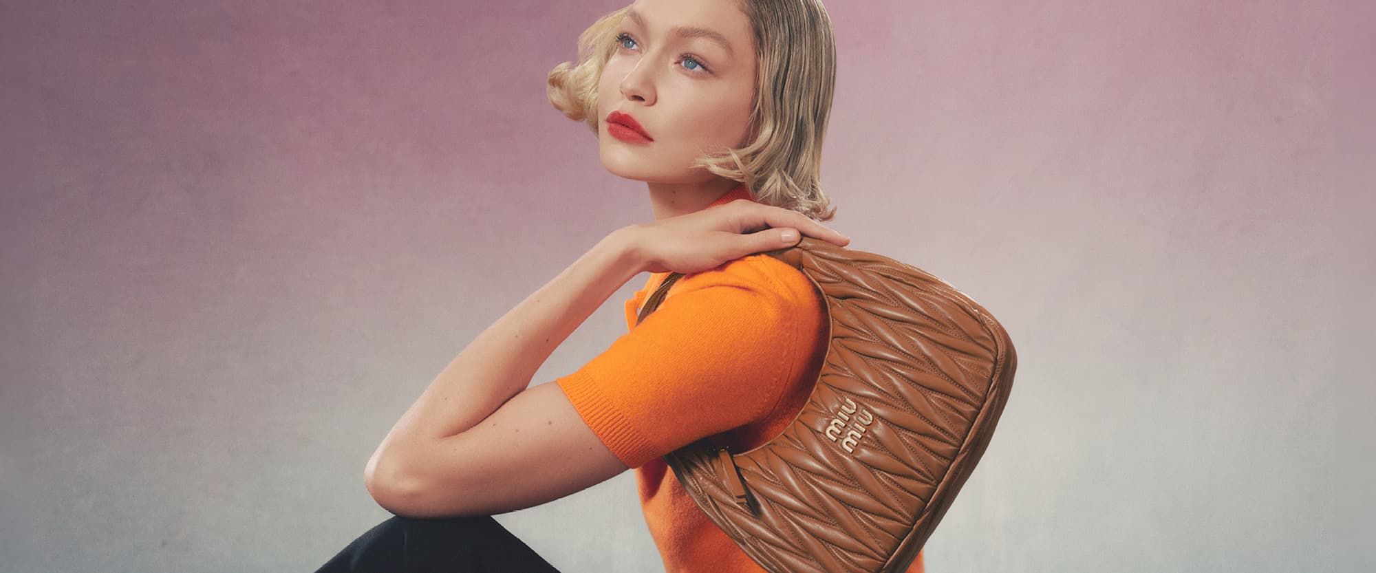 Fashion shot of a blond adult female model wearing an orange top holding a Miu Miu leather handbag in front of a pink gradient backdrop.