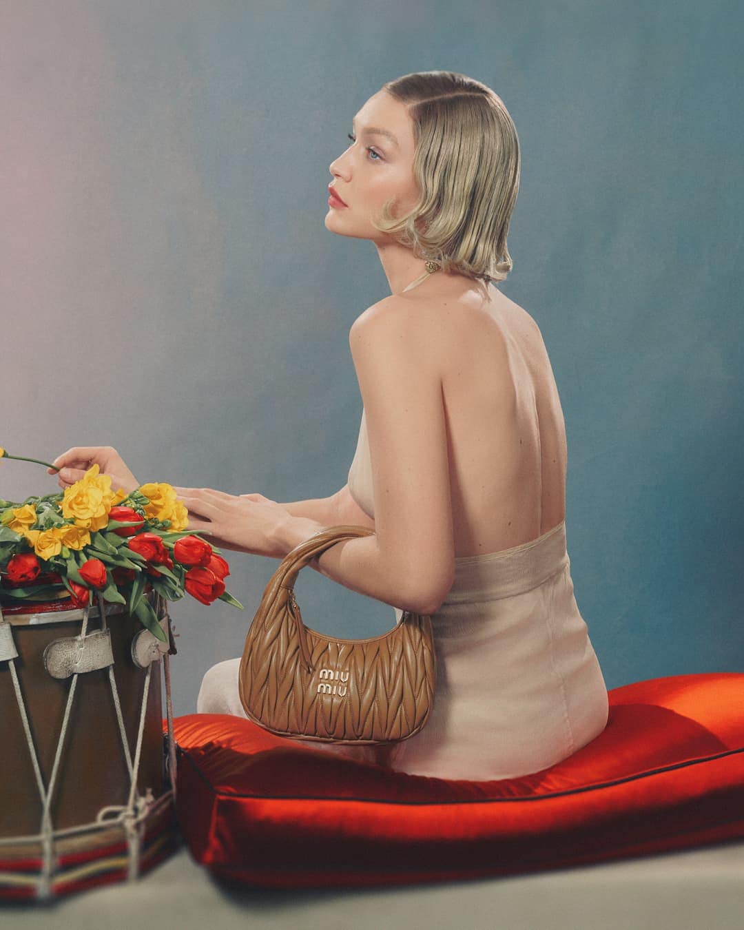 An adult female fashion model poses indoors, wearing a light dress and holding a brown Miu Miu handbag next to floral arrangement.
