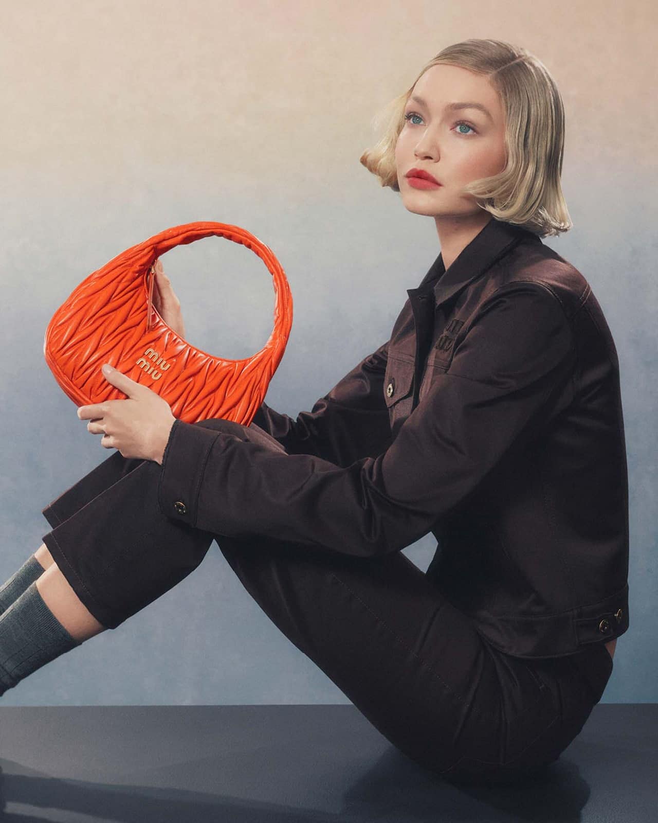 Gigi Hadid holding a bright orange quilted handbag while wearing a dark jacket against a textured gradient background