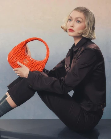 Gigi Hadid holding a bright orange quilted handbag while wearing a dark jacket against a textured gradient background