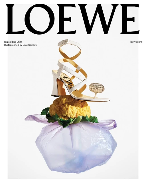 Loewe sandal with a white strap and embellishments is balanced atop yellow cauliflower and a light purple bag against a white background.