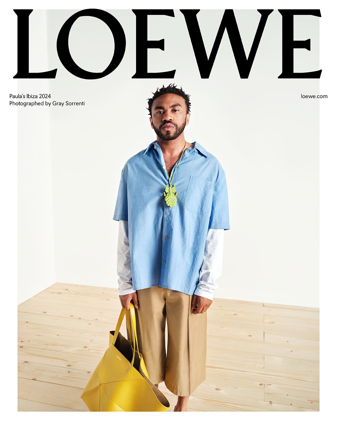 Fashion ad featuring a male model wearing a blue shirt, khaki pants, and holding a yellow bag, set against a simple white backdrop.