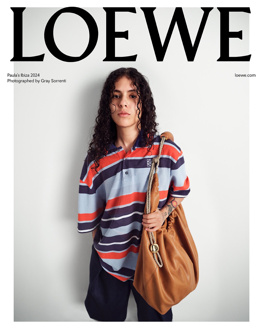 A model poses in front of a white wall wearing a striped polo shirt, dark pants, and a large tan leather bag slung over their shoulder.
