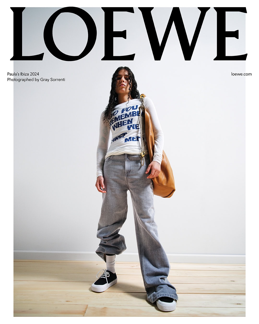 Fashion shot featuring a young woman in baggy jeans and a long-sleeve shirt, posing with a tan leather bag against a white backdrop.
