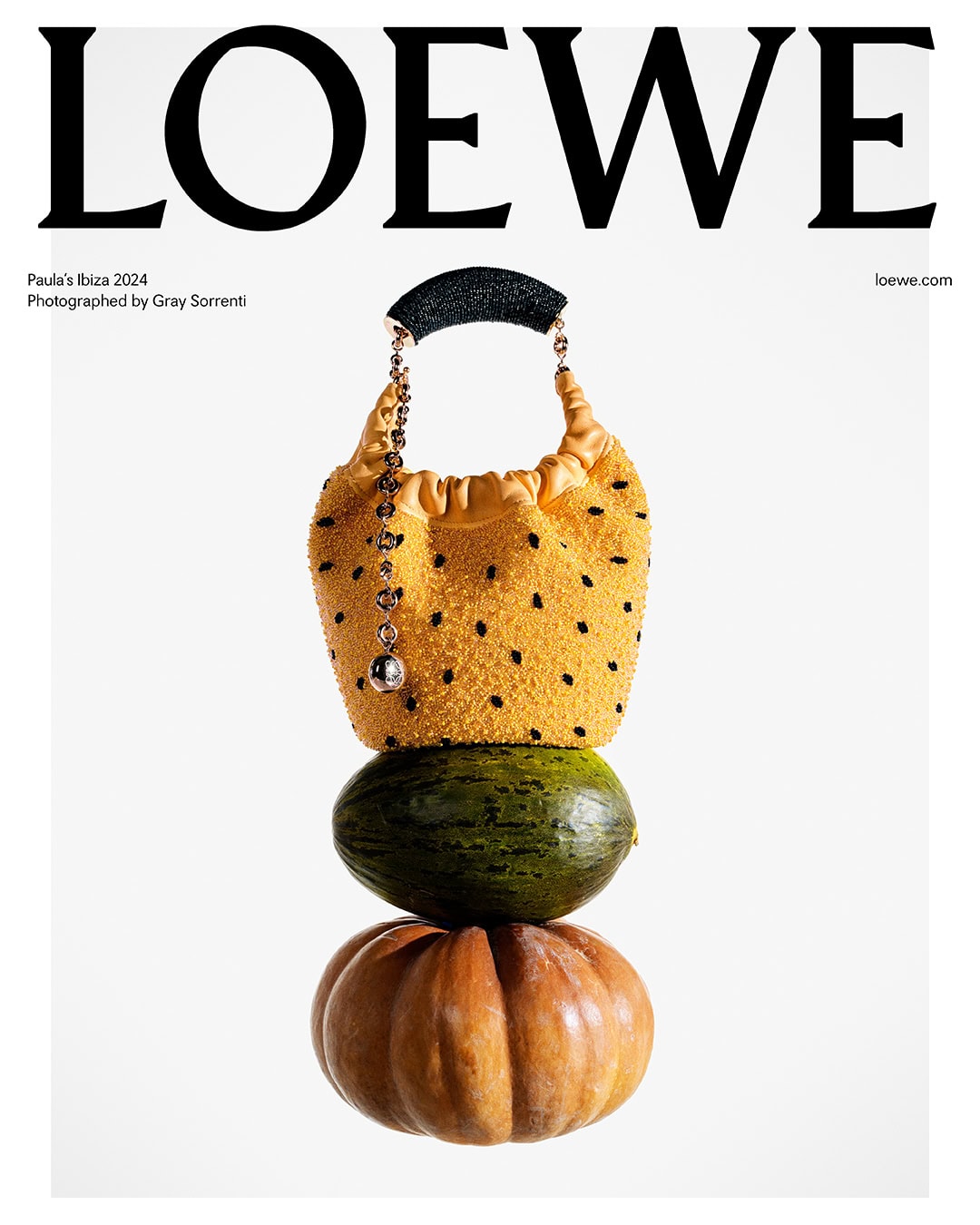 Still life of a Loewe handbag on top of a melon and pumpkin against a white background in a fashion product shot.