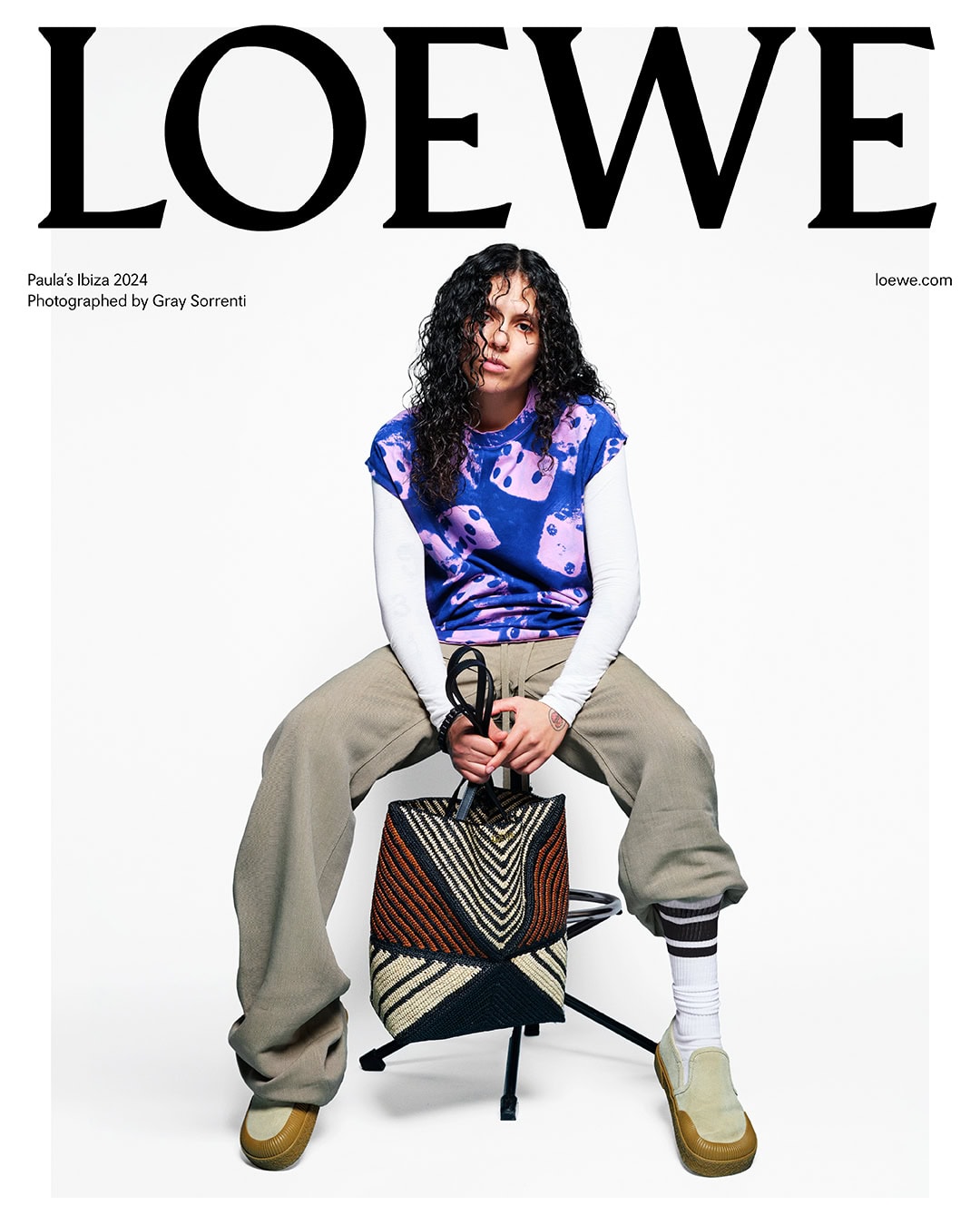 Fashion shot of person sitting on chair with geometric bag wearing colorful top, pants, socks, and loafers in a bright studio.