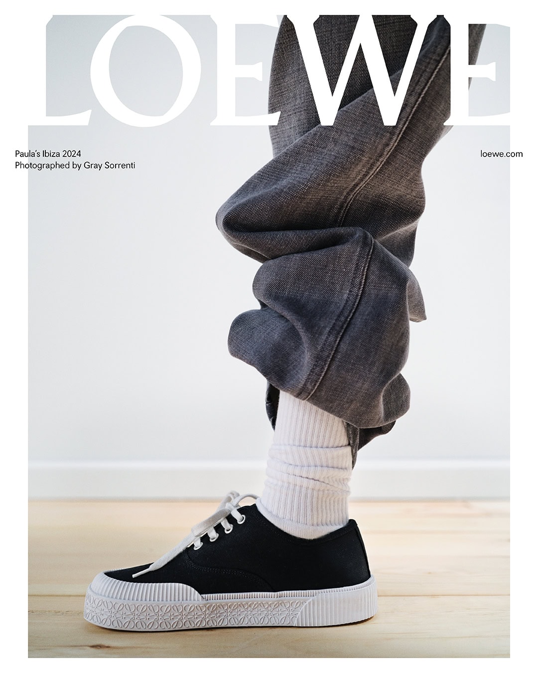 Fashion editorial featuring gray denim jeans, white socks, and black sneakers with white laces and soles on a light wood floor.