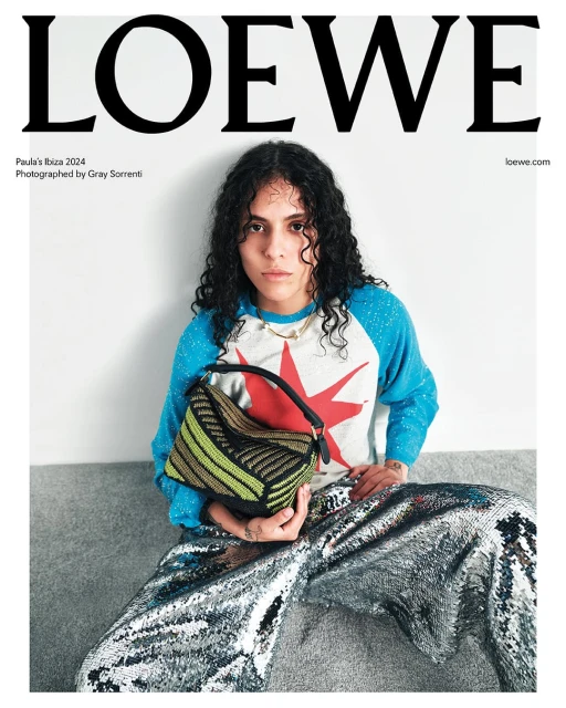 Magazine cover featuring a model wearing a patterned sweater and pants, seated and holding a striped handbag against a neutral background.