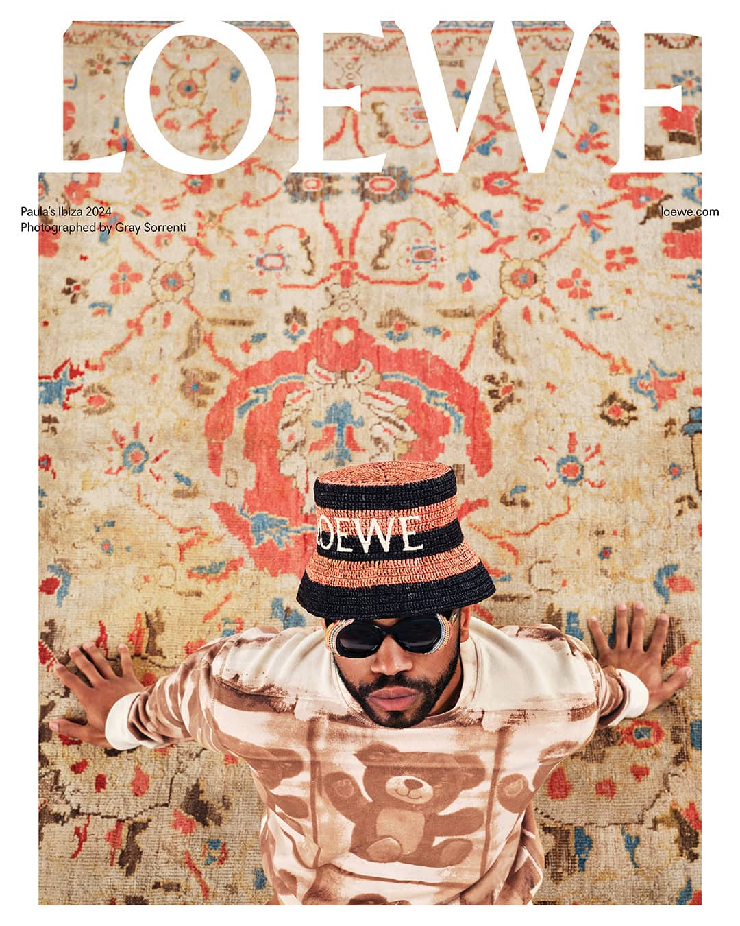 Magazine cover featuring a person lying on an ornate rug, wearing sunglasses, a Loewe hat, and a sweatshirt with a teddy bear design.