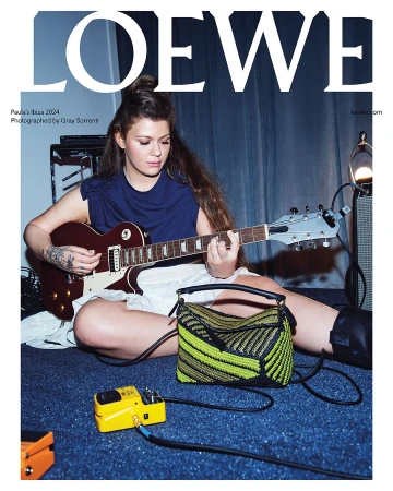 A fair-skinned woman with long brown hair plays an electric guitar while seated on the floor in a fashion editorial.