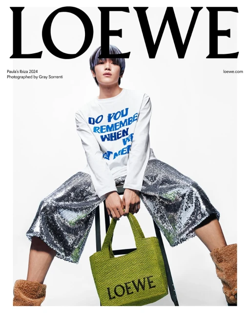 Fashion editorial shot of a model wearing a white long sleeve shirt, sequin pants, and brown boots, holding a green woven tote bag.