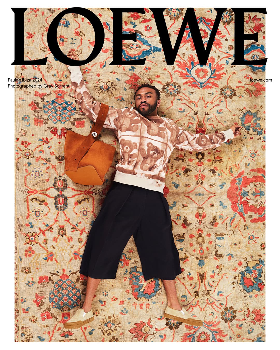 A man wearing a teddy bear print sweater and black culottes lies spread-eagle on a large, intricately patterned floral rug with a suede bag.