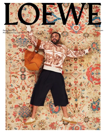 A man wearing a teddy bear print sweater and black culottes lies spread-eagle on a large, intricately patterned floral rug with a suede bag.